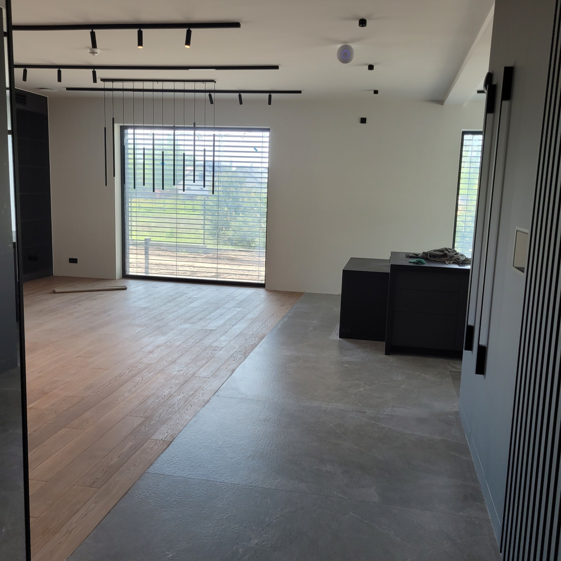 Nowoczesne wnętrze z parkietem ułożonym na części podłogi, reszta wyłożona szarymi płytkami. Okno z żaluzjami i czarne meble dopełniają minimalistyczny wystrój.