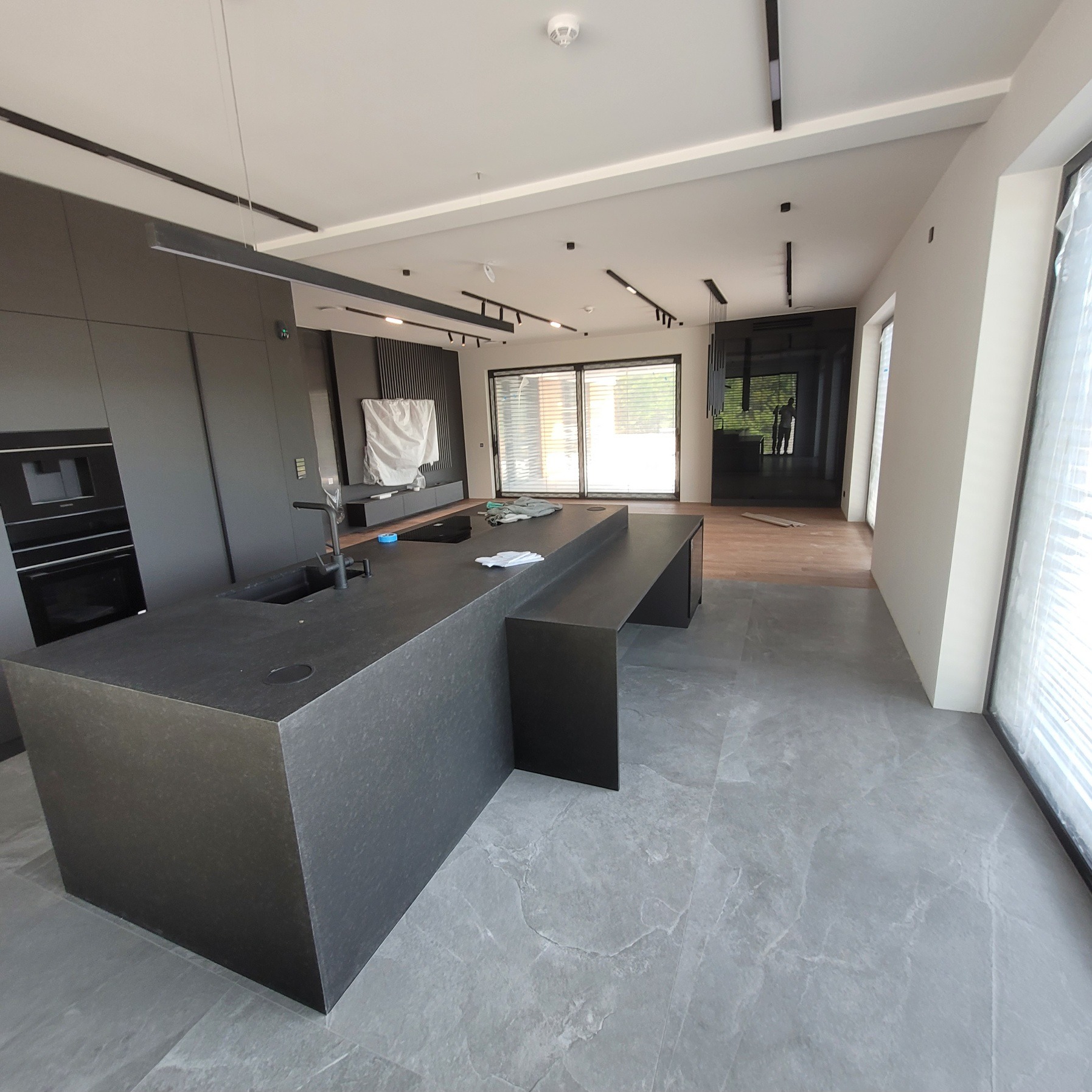 Nowoczesna, szara kuchnia z wyspą i marmurową podłogą. Minimalistyczny design, widoczne oświetlenie sufitowe. Częściowo zasłonięte okno.