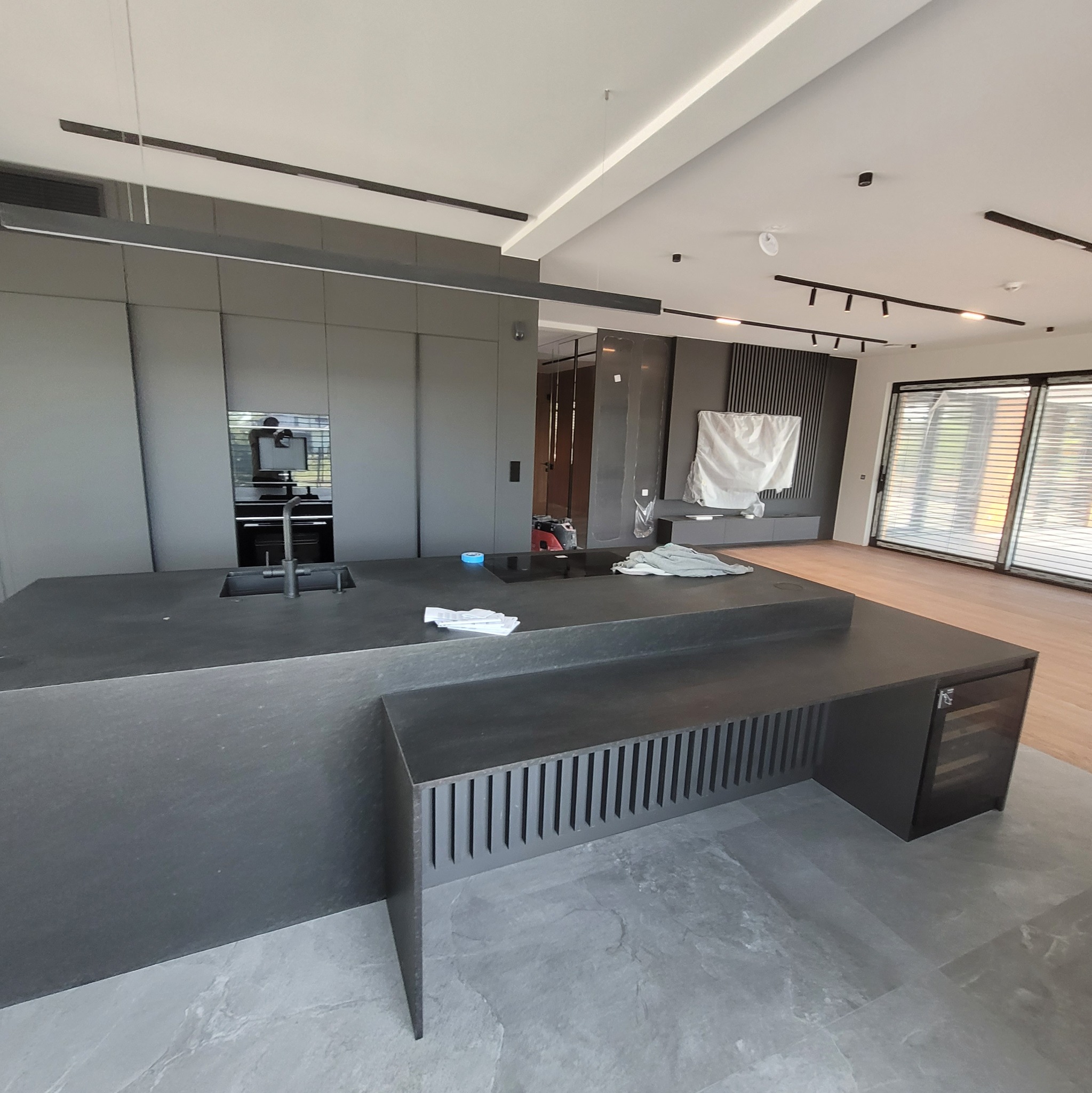 Nowoczesna kuchnia w odcieniach szarości z wyspą. Widoczne minimalistyczne szafki, oświetlenie sufitowe i duże okno z żaluzjami. Podłoga wyłożona szarymi płytkami.