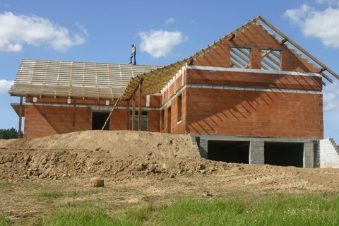 Wznoszenie dwukondygnacyjnego domu z czerwonej cegły ceramicznej; widoczna konstrukcja dachu z łatami oraz pracownik na dachu, teren wokół domu nierówny, z nasypami ziemi.