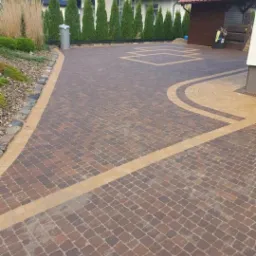 Ułożona kostka brukowa w odcieniach brązu i beżu tworząca wzór na podjeździe przed domem z tujami i ozdobnymi trawami w tle.