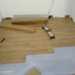 Układanie paneli podłogowych w trakcie pracy: widoczna warstwa izolacyjna, ułożone panele, narzędzia do cięcia i dobijania paneli, oraz zapas paneli przy ścianie.