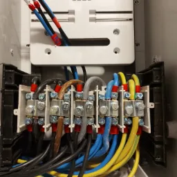 Rozdzielnica elektryczna z podłączonymi przewodami o różnych kolorach (czarny, niebieski, brązowy, szary, żółto-zielony) do listew zaciskowych, widoczne śruby i oznaczenia na przewodach.