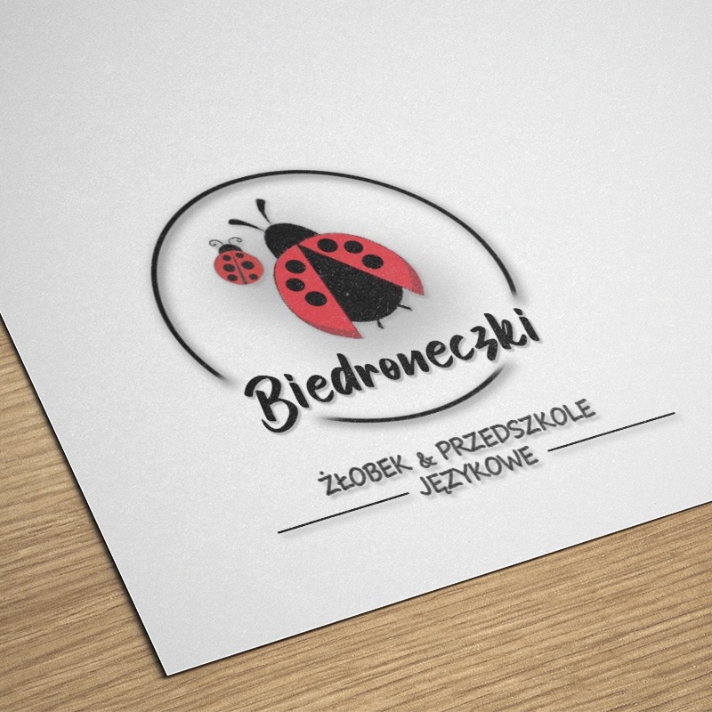 Logo żłobka i przedszkola językowego 'Biedroneczki' z motywem dwóch biedronek w czarno-czerwonej kolorystyce, umieszczone na białym papierze z delikatną fakturą
