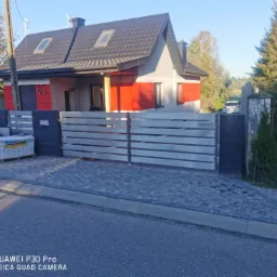 Nowoczesna brama wjazdowa z poziomych, metalowych profili, zamontowana przed domem z czerwono-szarą elewacją, widoczne kostki brukowe na paletach i podjazd z kostki.