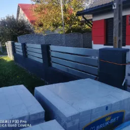 Nowoczesne ogrodzenie posesji z poziomych, metalowych profili w kolorze srebrnym, z ciemnoszarymi słupkami betonowymi, częściowo ukończone; na pierwszym planie kostka brukowa w foliowych opakowaniach.