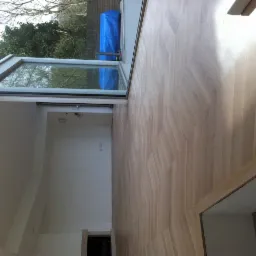 Jasny parkiet ułożony w jodełkę w nowym, pustym pomieszczeniu z dużym oknem dachowym i widokiem na drzewa.