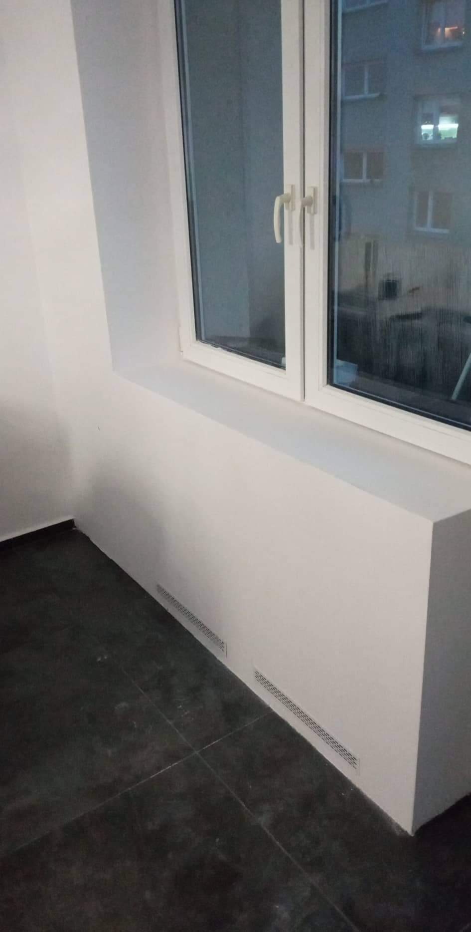 Białe okno PCV z parapetem zabudowanym płytą kartonowo-gipsową z kratkami wentylacyjnymi, ciemna podłoga z płytek, widok na budynek za oknem.