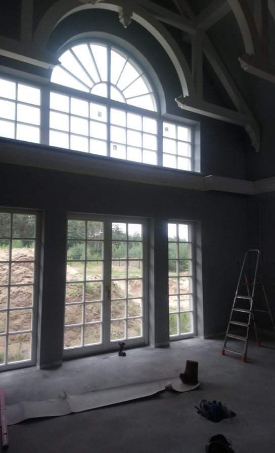 Wnętrze w trakcie prac wykończeniowych: duże okna z widokiem na zewnątrz, drabina oparta o ścianę, rozłożona folia ochronna na podłodze, widoczne narzędzia.