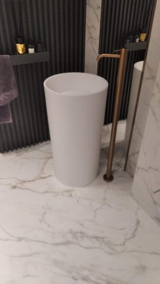 Nowoczesna, wolnostojąca, biała umywalka cylindryczna z mosiężną baterią, na marmurowej podłodze, z elementami dekoracyjnymi na ścianie w tle.