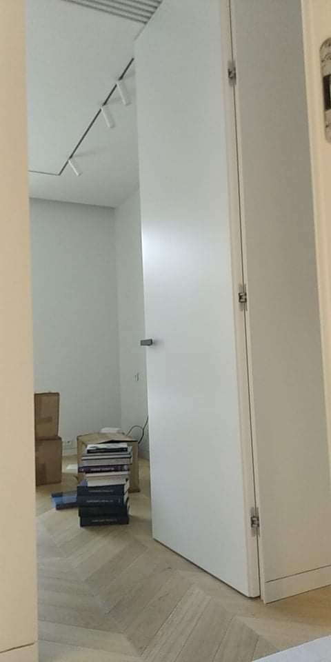 Wnętrze pokoju z ułożoną podłogą w jodełkę, białe ściany, minimalistyczne oświetlenie sufitowe, białe drzwi z widocznymi zawiasami i sterta książek w kartonach, sugerująca przeprowadzkę lub etap...