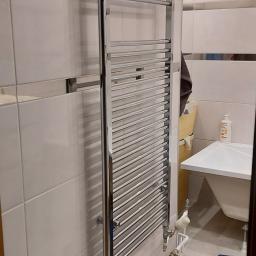 Hydra-luk - Chromowany grzejnik drabinkowy zamontowany na białej, kafelkowanej ścianie łazienki, widoczne rury doprowadzające wodę i niebieska skrzynka narzędziowa na podłodze.