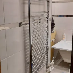 Chromowany grzejnik drabinkowy zamontowany na białej, kafelkowanej ścianie łazienki, widoczne rury doprowadzające wodę i niebieska skrzynka narzędziowa na podłodze.