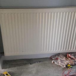 Hydra-luk - Biały, żebrowany grzejnik panelowy zamontowany na szarej ścianie, widoczne narzędzia i szmata na podłodze w trakcie prac instalacyjnych.