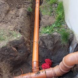 Hydra-luk - Pomarańczowe rury kanalizacyjne ułożone w wykopie ziemnym wzdłuż ściany budynku, widoczny poziomica i fragmenty potłuczonej rury.