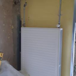 Hydra-luk - Nowy, biały grzejnik panelowy zamontowany na żółtej ścianie w trakcie prac instalacyjnych, widoczne rury doprowadzające, fragment ściany w stanie surowym i poziomica.