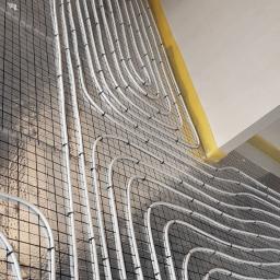 Hydra-luk - Montaż ogrzewania podłogowego: białe rury grzewcze rozłożone na srebrnej folii izolacyjnej z czarną siatką, przymocowane czarnymi klipsami, widoczne żółte taśmy na styku ściany i podłogi.