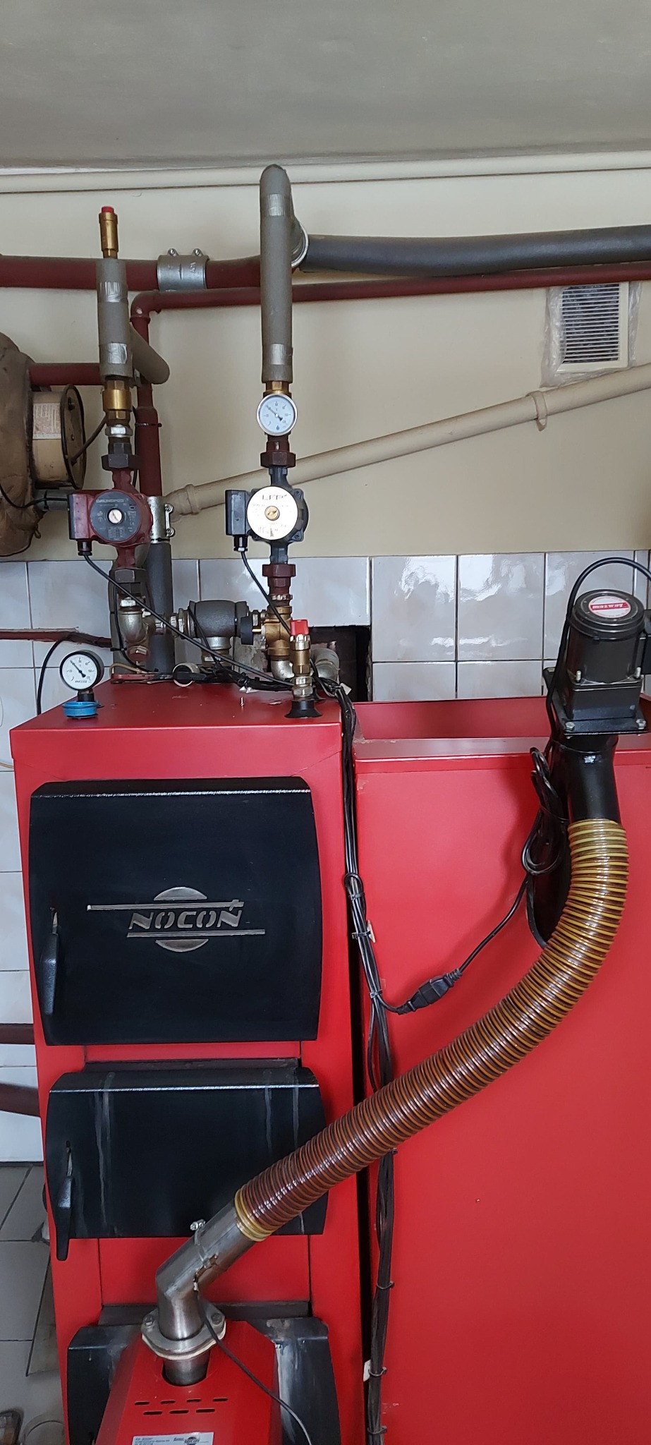Czerwony piec CO marki Nocon z podłączonymi rurami i manometrami w kotłowni z białymi kafelkami.
