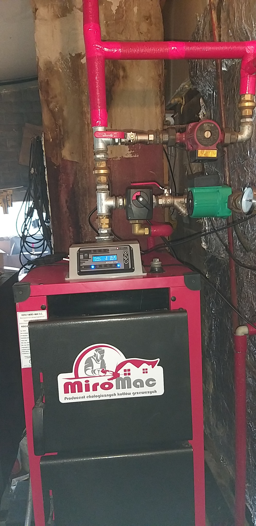 Czerwono-czarny kocioł grzewczy marki MiroMac z podłączonym sterownikiem Techmix Argos PID i rurami w izolacji w kolorze fuksji, widoczne zawory, pompy obiegowe oraz manometr.