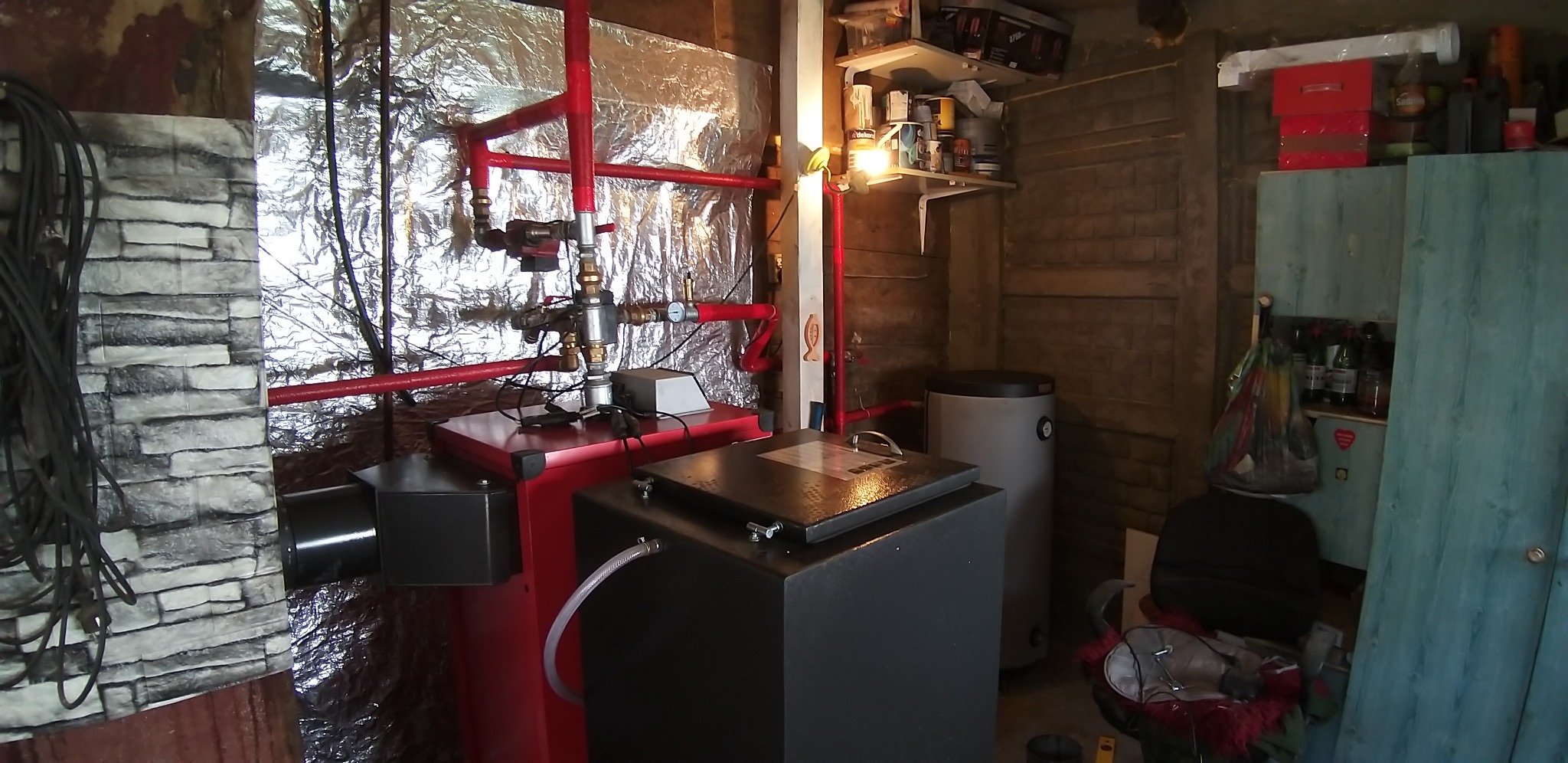 Nowoczesny piec na paliwo stałe z czerwonymi rurami w kotłowni, widoczne elementy instalacji, izolacja folią aluminiową na ścianie i półki z narzędziami.
