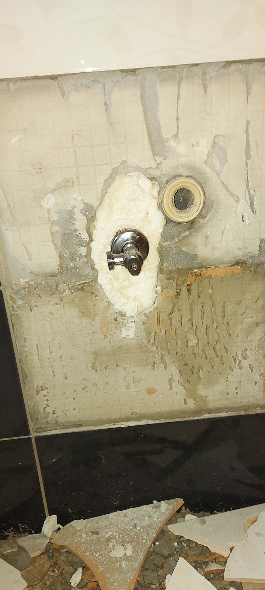 Niedokończona ściana w łazience z zamontowanym, chromowanym zaworem i fragmentem rury, otoczonymi pianką montażową; widoczne fragmenty starej glazury i gruz po remoncie.