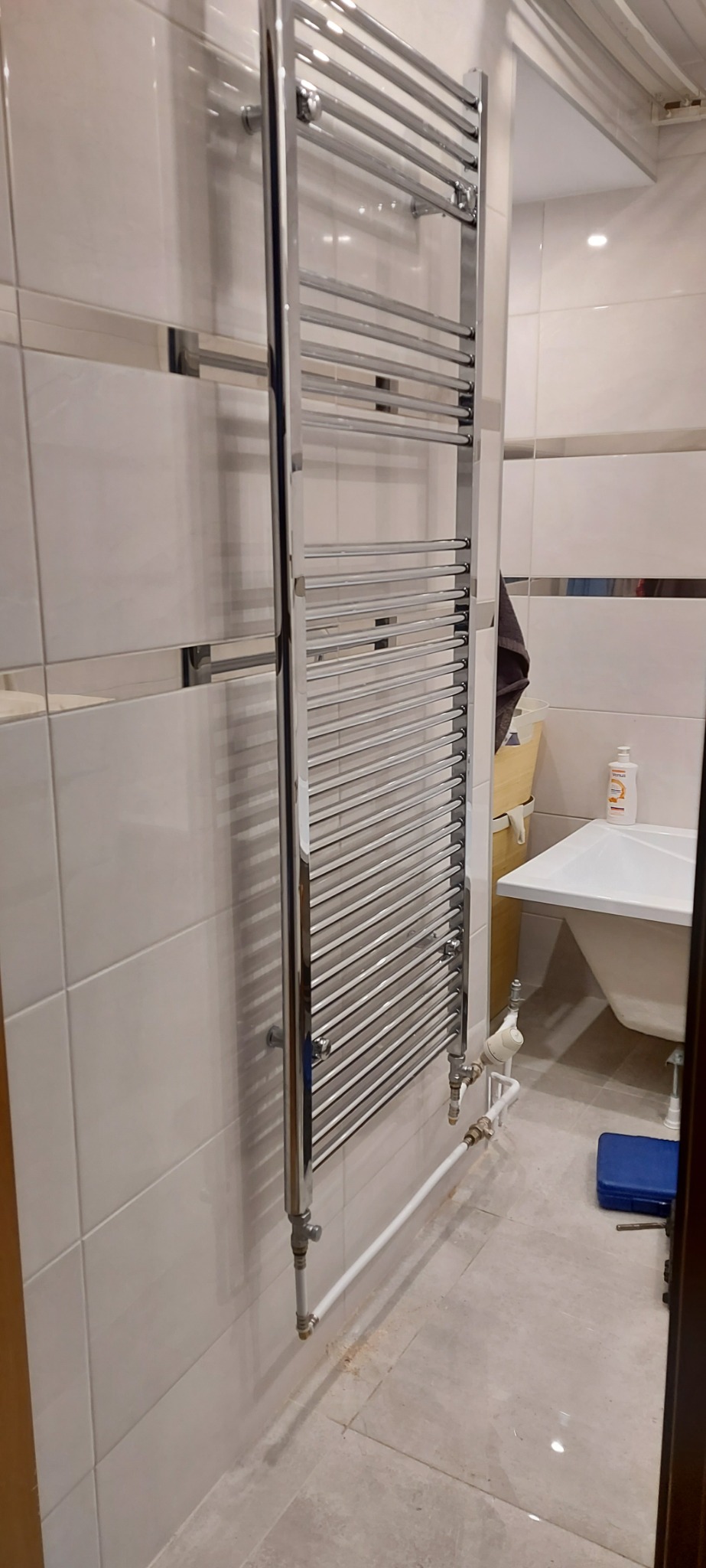 Chromowany grzejnik drabinkowy zamontowany na białej, kafelkowanej ścianie łazienki, widoczne rury doprowadzające wodę i niebieska skrzynka narzędziowa na podłodze.