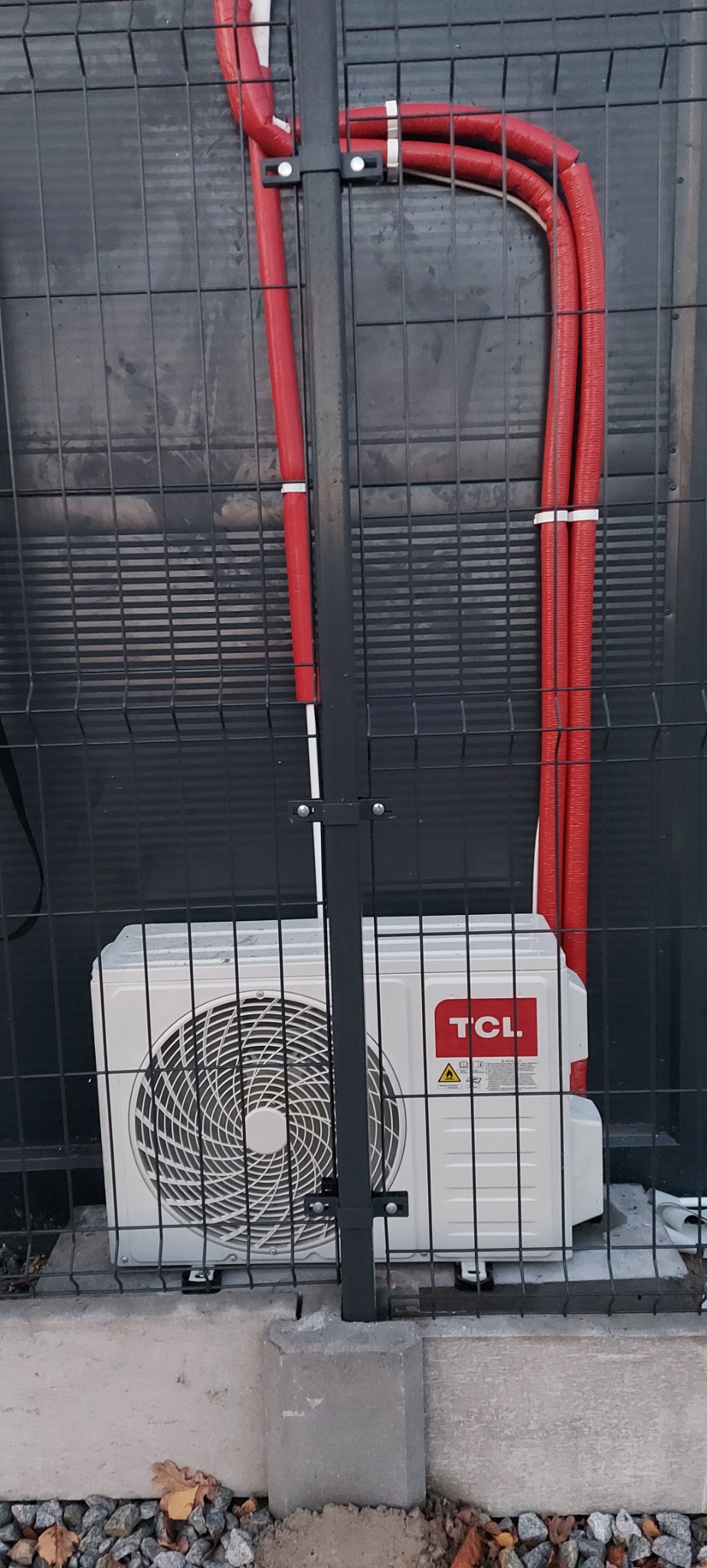 Biały klimatyzator TCL zamontowany przy metalowym ogrodzeniu, z widocznymi czerwonymi rurami instalacyjnymi zabezpieczonymi białymi opaskami.