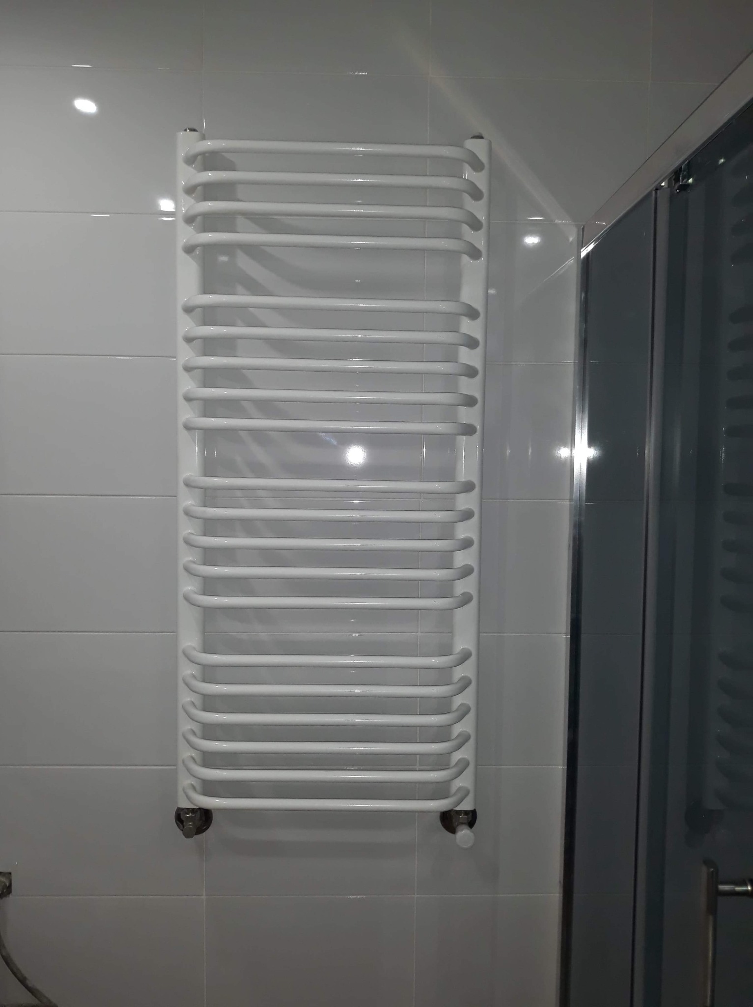 Biały, drabinkowy grzejnik łazienkowy zamontowany na białych, błyszczących płytkach, obok kabiny prysznicowej z przezroczystymi drzwiami.