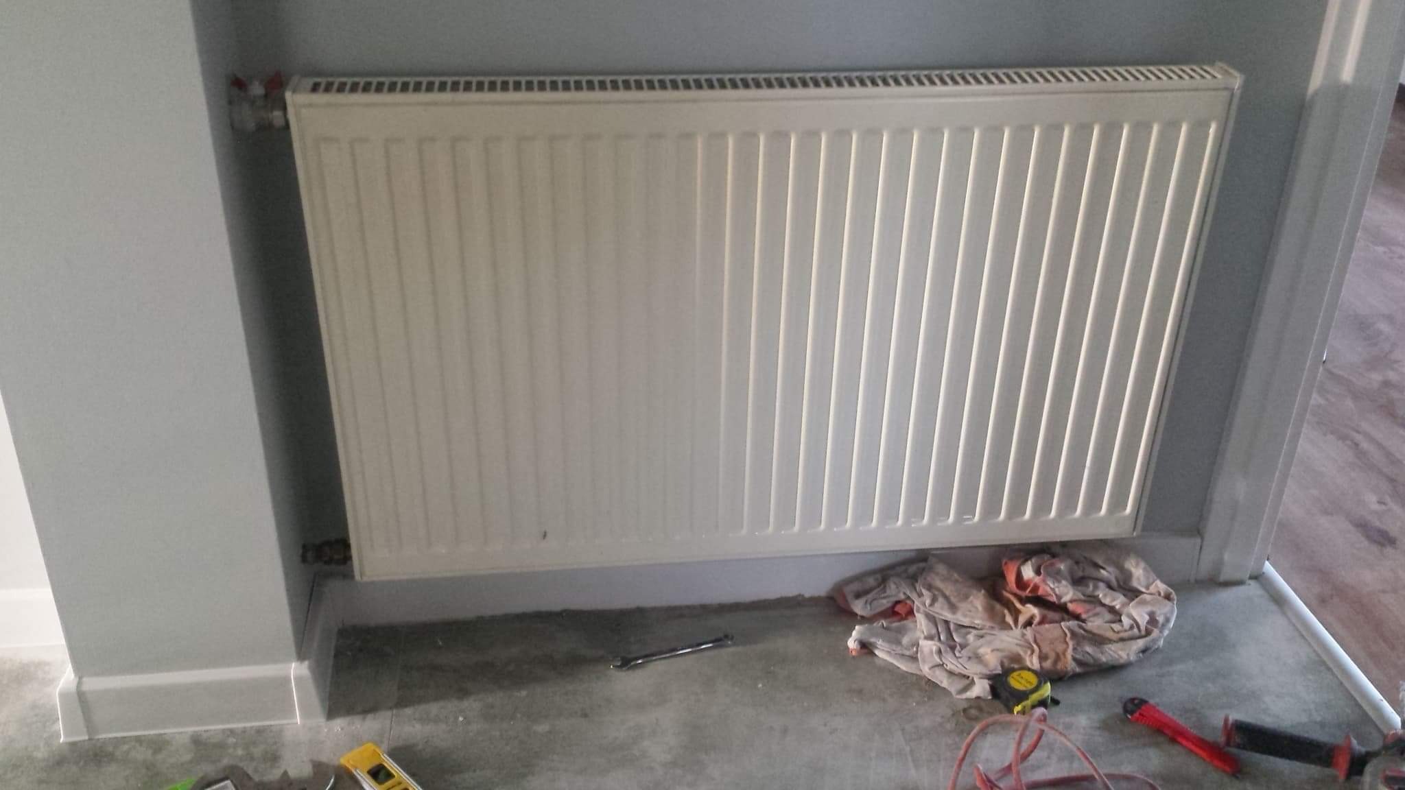 Biały, żebrowany grzejnik panelowy zamontowany na szarej ścianie, widoczne narzędzia i szmata na podłodze w trakcie prac instalacyjnych.