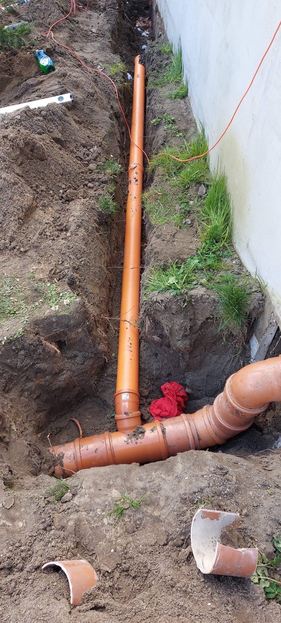 Pomarańczowe rury kanalizacyjne ułożone w wykopie ziemnym wzdłuż ściany budynku, widoczny poziomica i fragmenty potłuczonej rury.