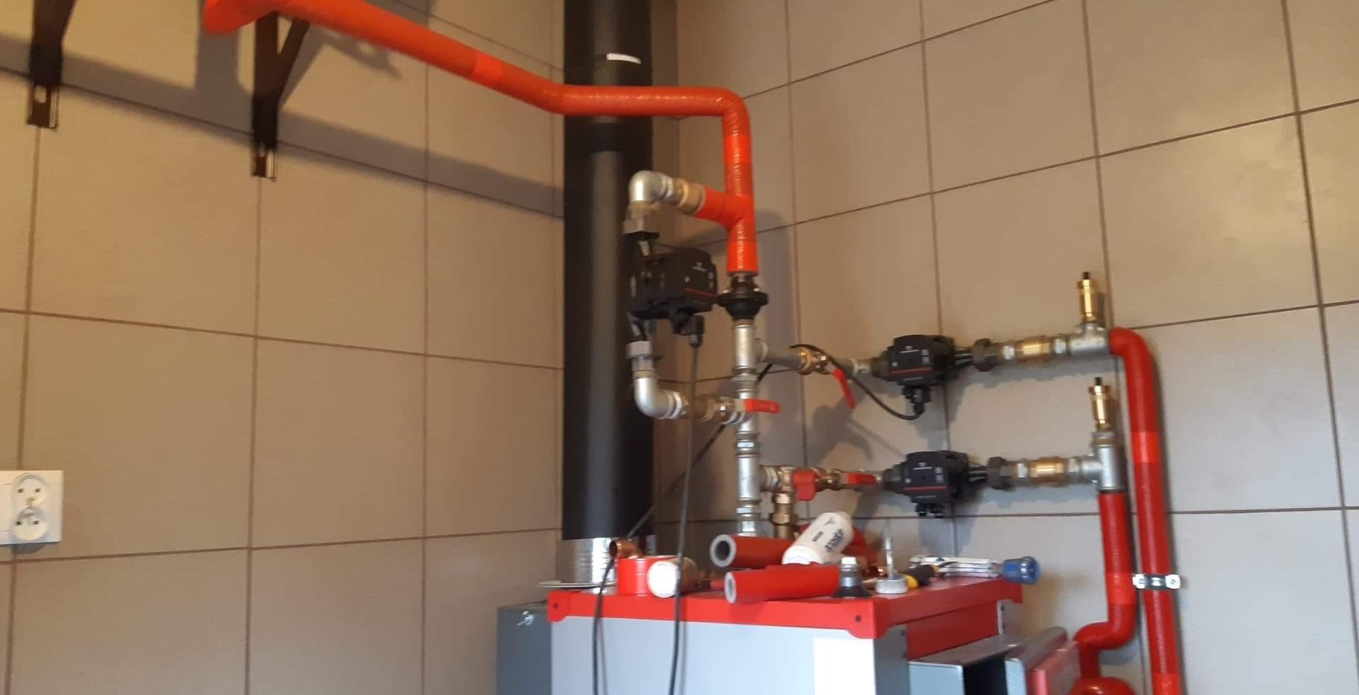 Instalacja grzewcza z piecem, rurami w izolacji termicznej i pompami obiegowymi na tle kafelkowanej ściany. Widoczne narzędzia i materiały montażowe na piecu.
