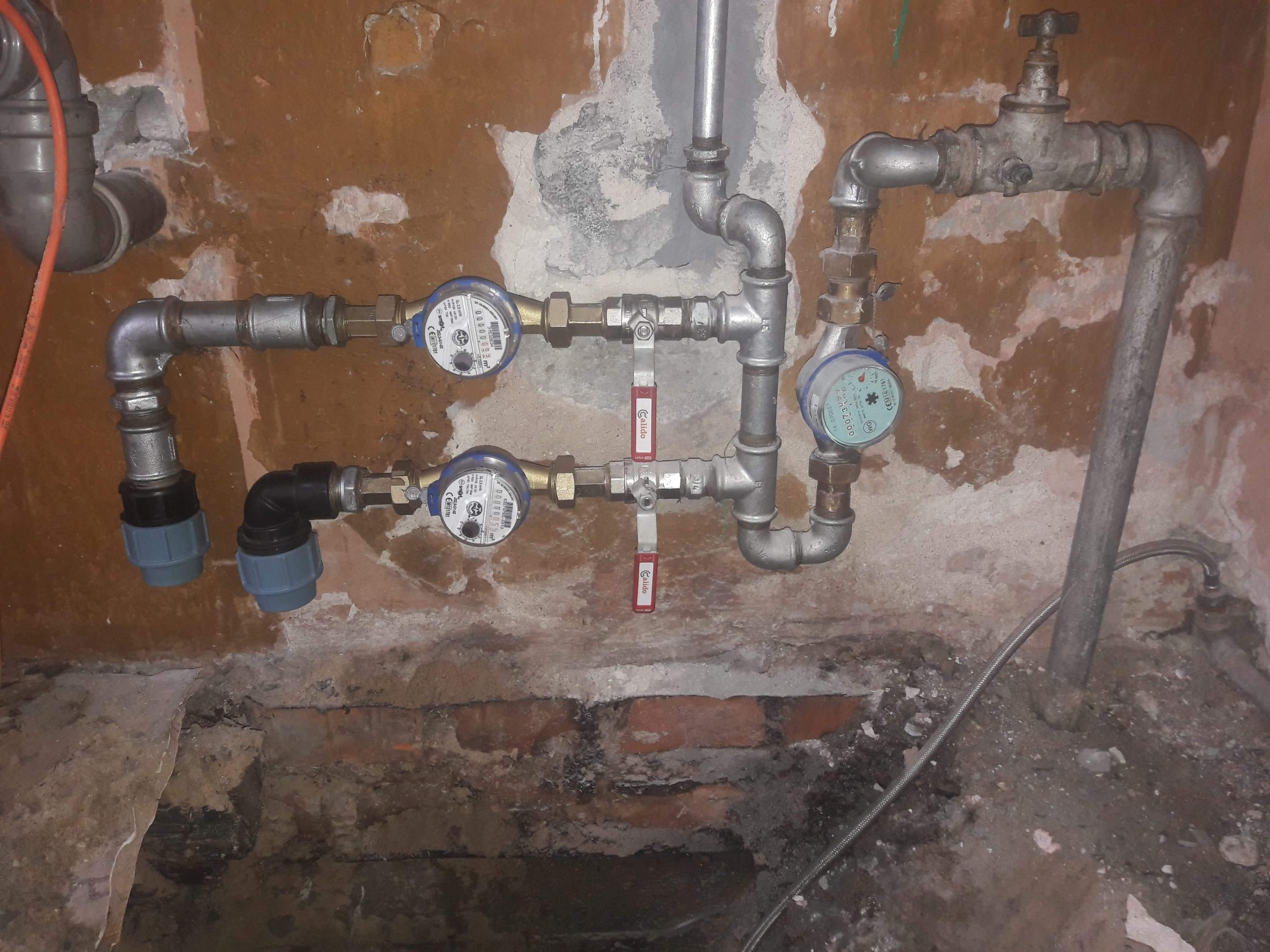 Instalacja wodna z dwoma wodomierzami i zaworami odcinającymi na tle surowej ściany z widocznymi śladami po remoncie i fragmentami cegieł.