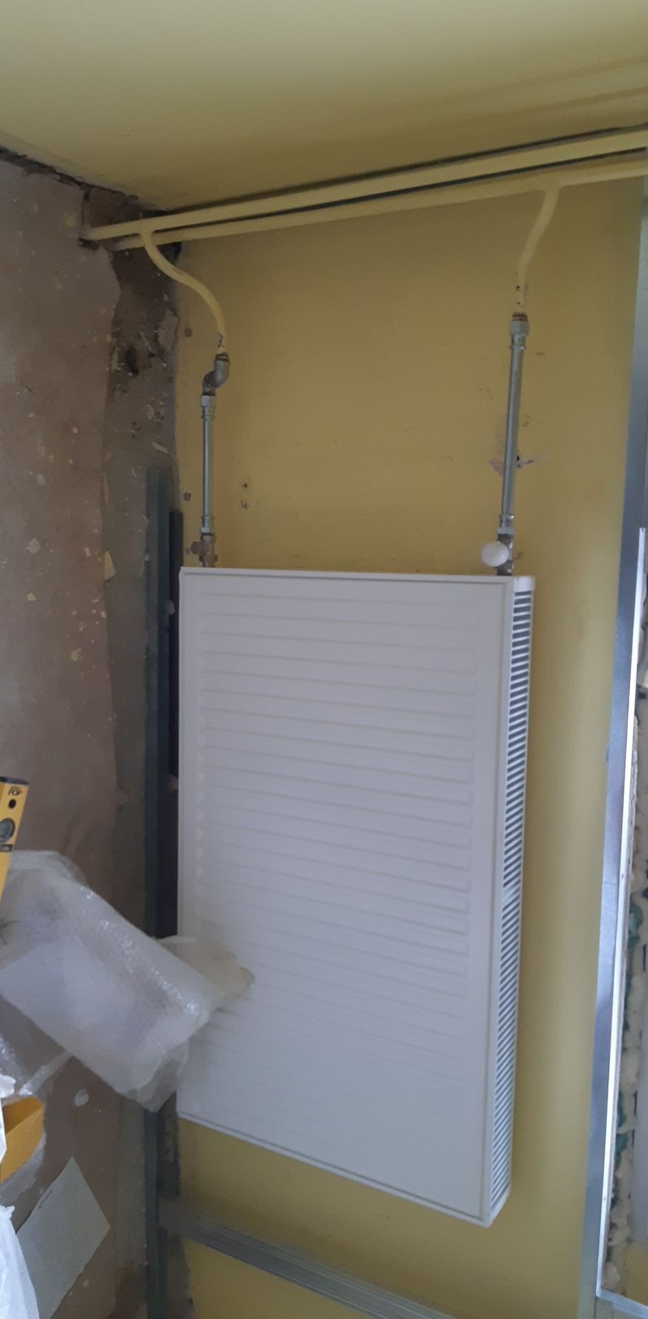 Nowy, biały grzejnik panelowy zamontowany na żółtej ścianie w trakcie prac instalacyjnych, widoczne rury doprowadzające, fragment ściany w stanie surowym i poziomica.