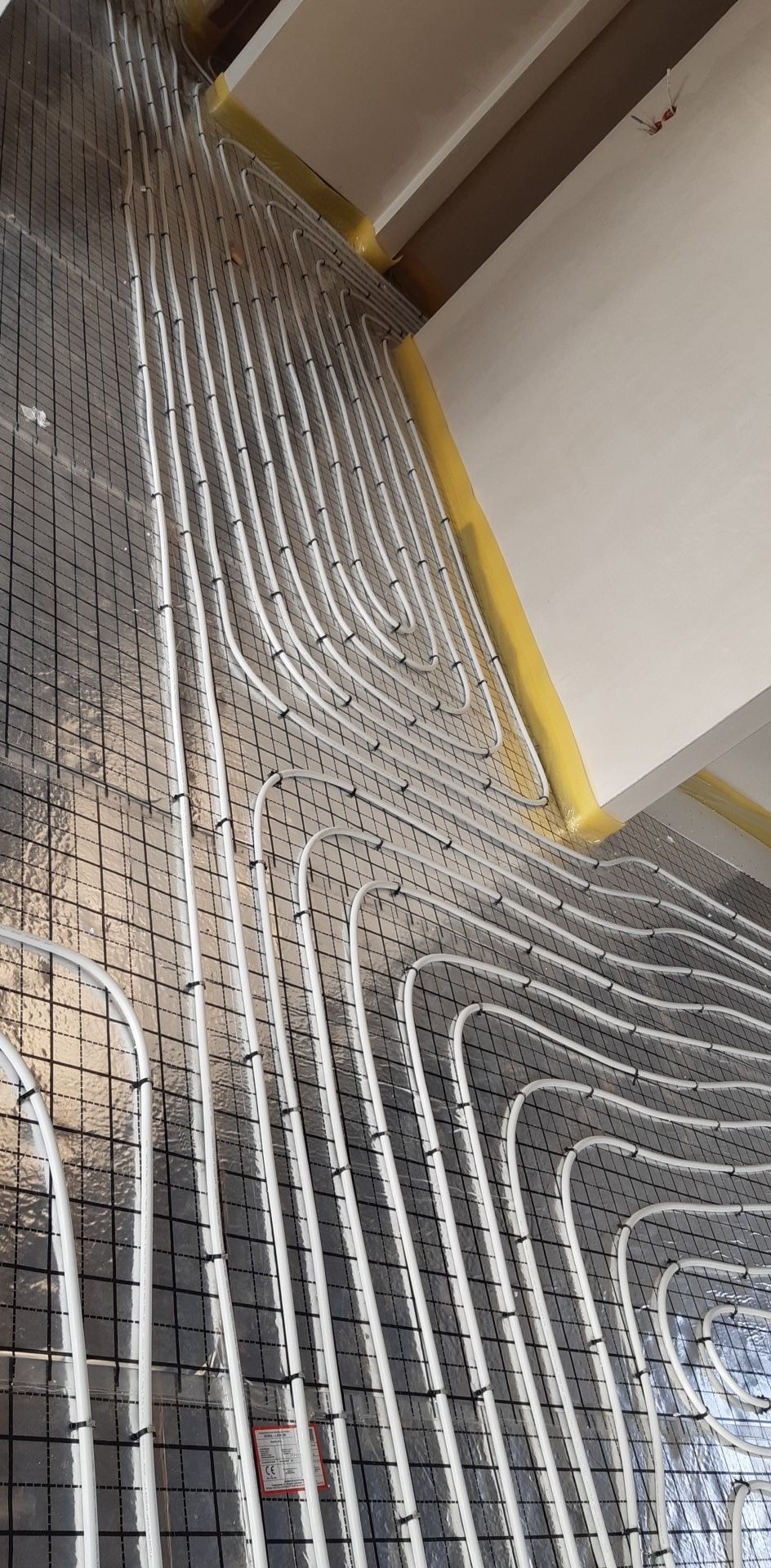 Montaż ogrzewania podłogowego: białe rury grzewcze rozłożone na srebrnej folii izolacyjnej z czarną siatką, przymocowane czarnymi klipsami, widoczne żółte taśmy na styku ściany i podłogi.