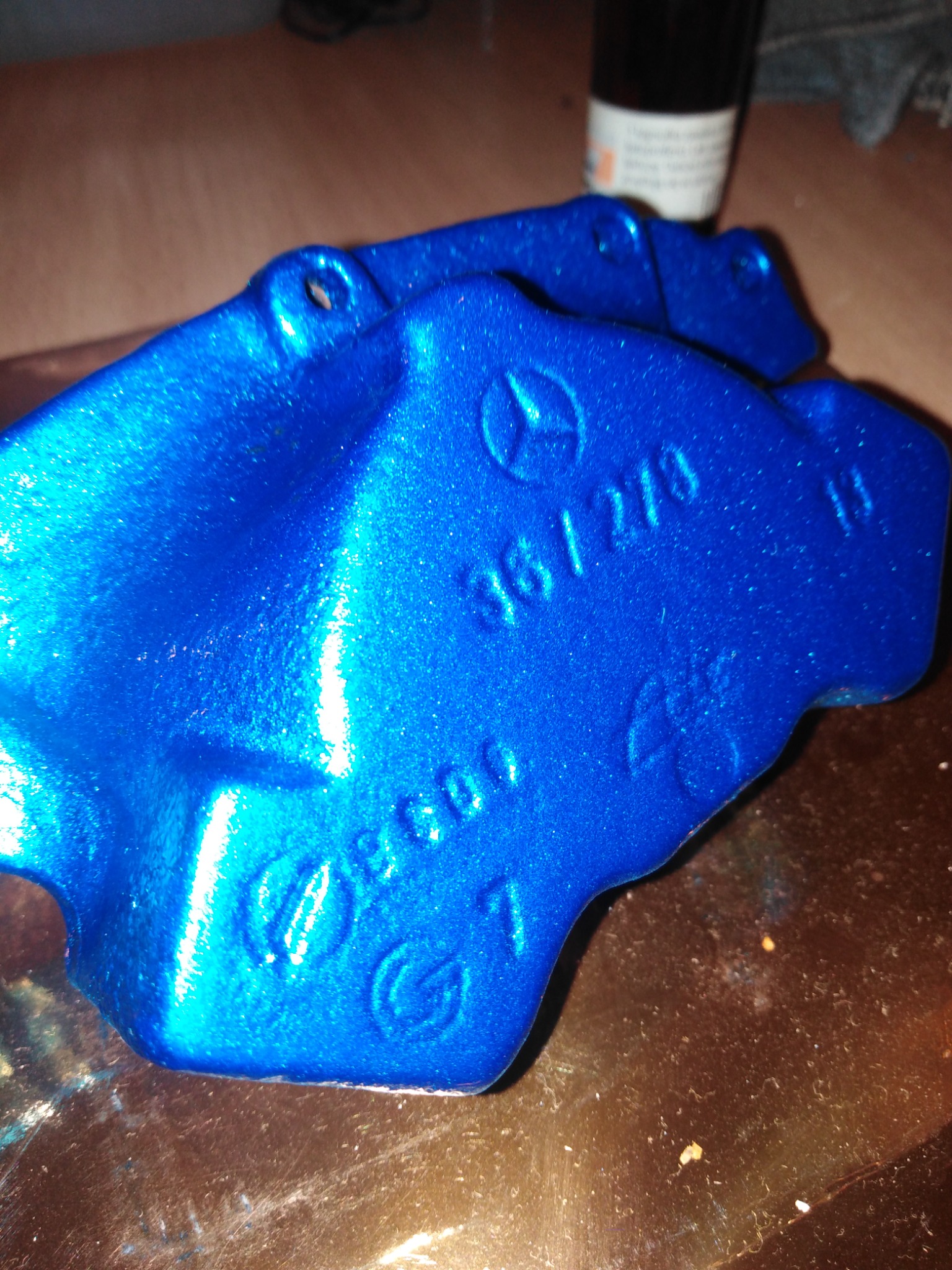 Błyszczący, niebieski zacisk hamulcowy Mercedes-Benz po malowaniu proszkowym, widoczne oznaczenia modelu i logo, leżący na brązowej powierzchni.