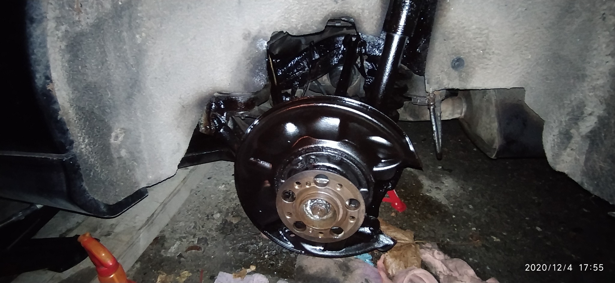 Element zawieszenia samochodu po renowacji, pokryty czarną, błyszczącą farbą, widoczny zacisk hamulcowy i przewody. Zdjęcie wykonane w warsztacie, widoczne zabrudzenia i narzędzia.