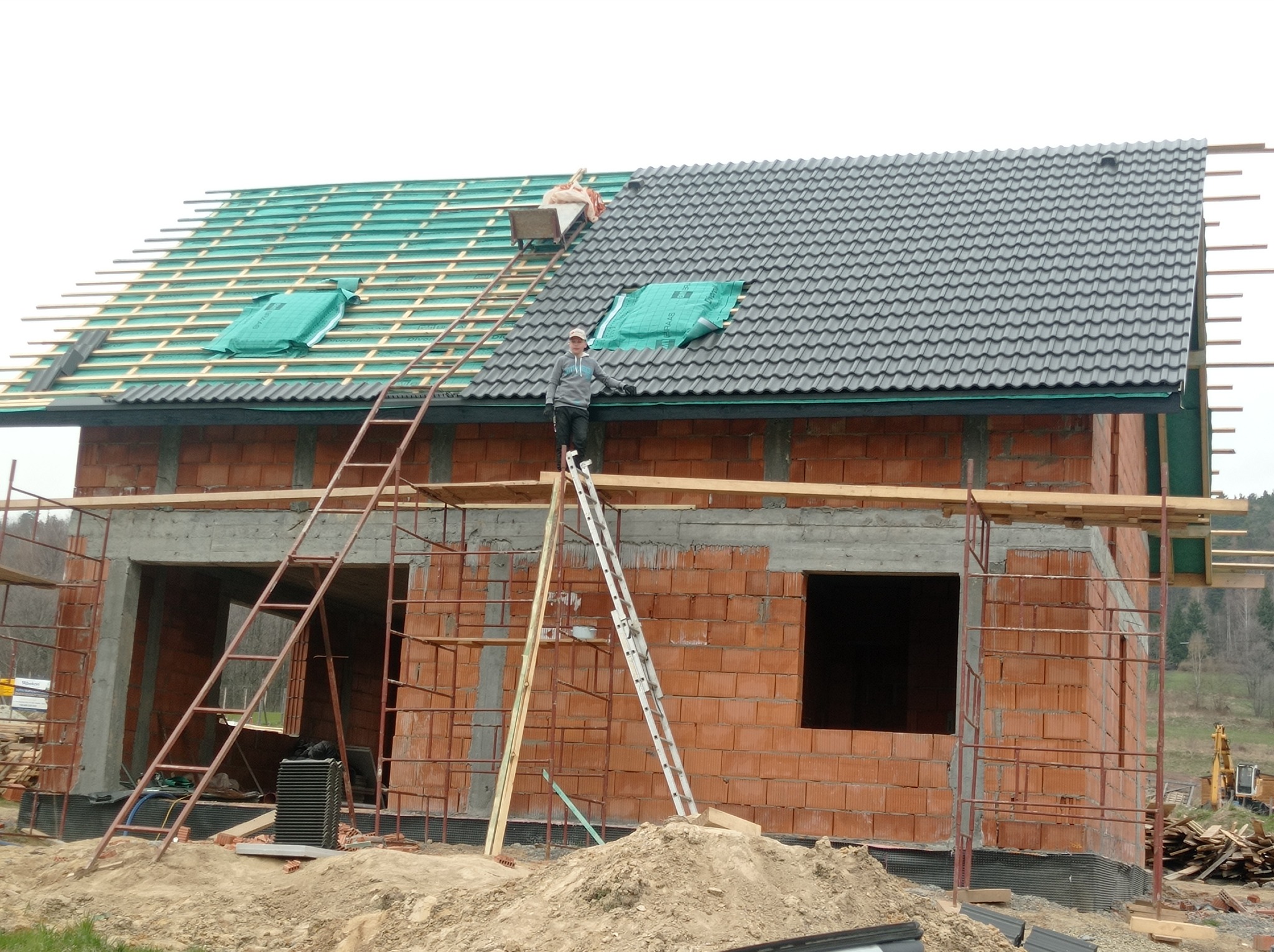 Budowa domu murowanego: robotnicy montują dachówkę na częściowo pokrytym zieloną membraną dachu, widoczne rusztowania i stos cegieł