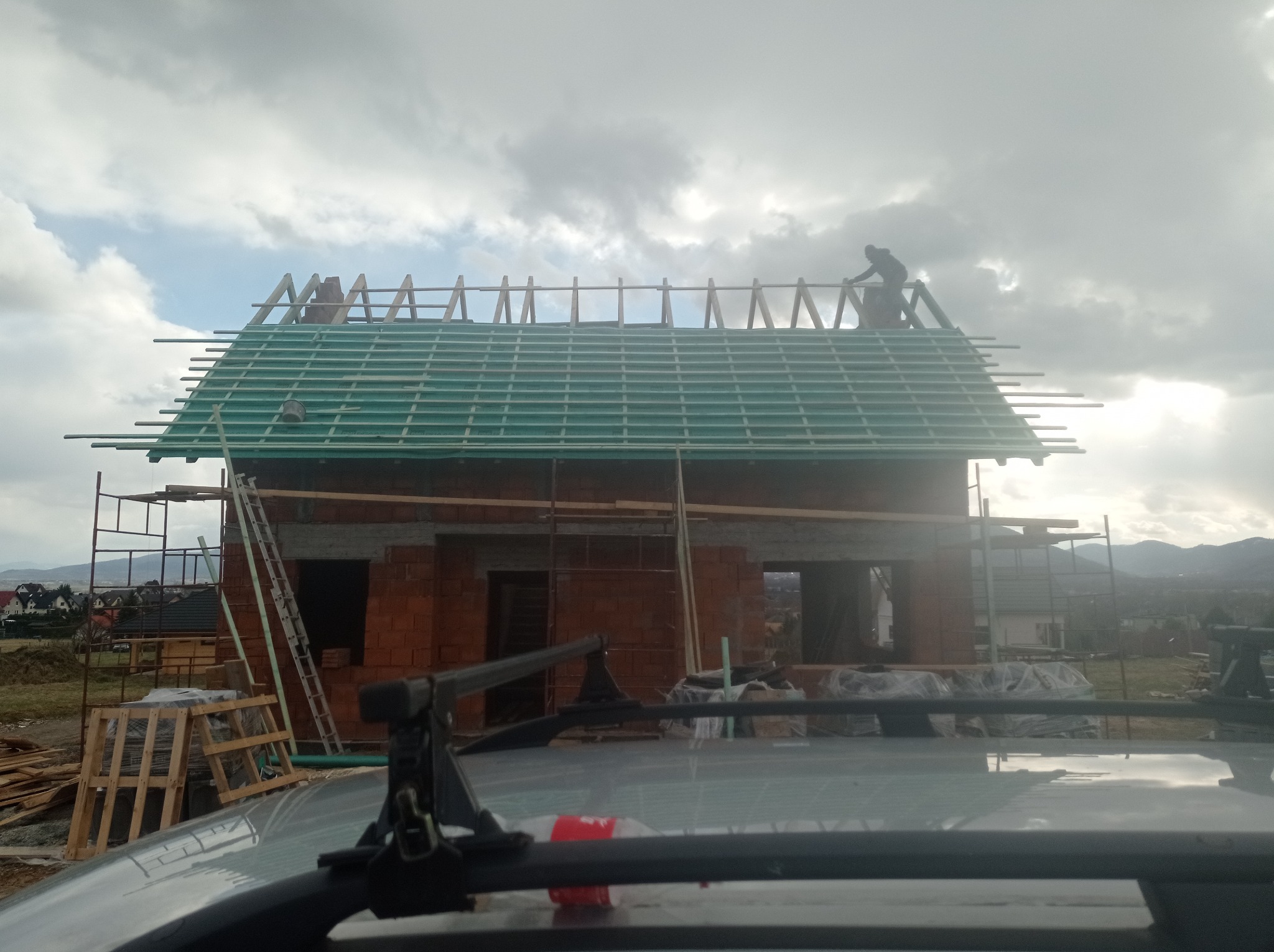 Budowa domu murowanego w trakcie: widok z dachu samochodu na wznoszoną konstrukcję z czerwonej cegły, z zielonymi łatami dachowymi i pracującym dekarzem na szczycie dachu, na tle pochmurnego nieba...