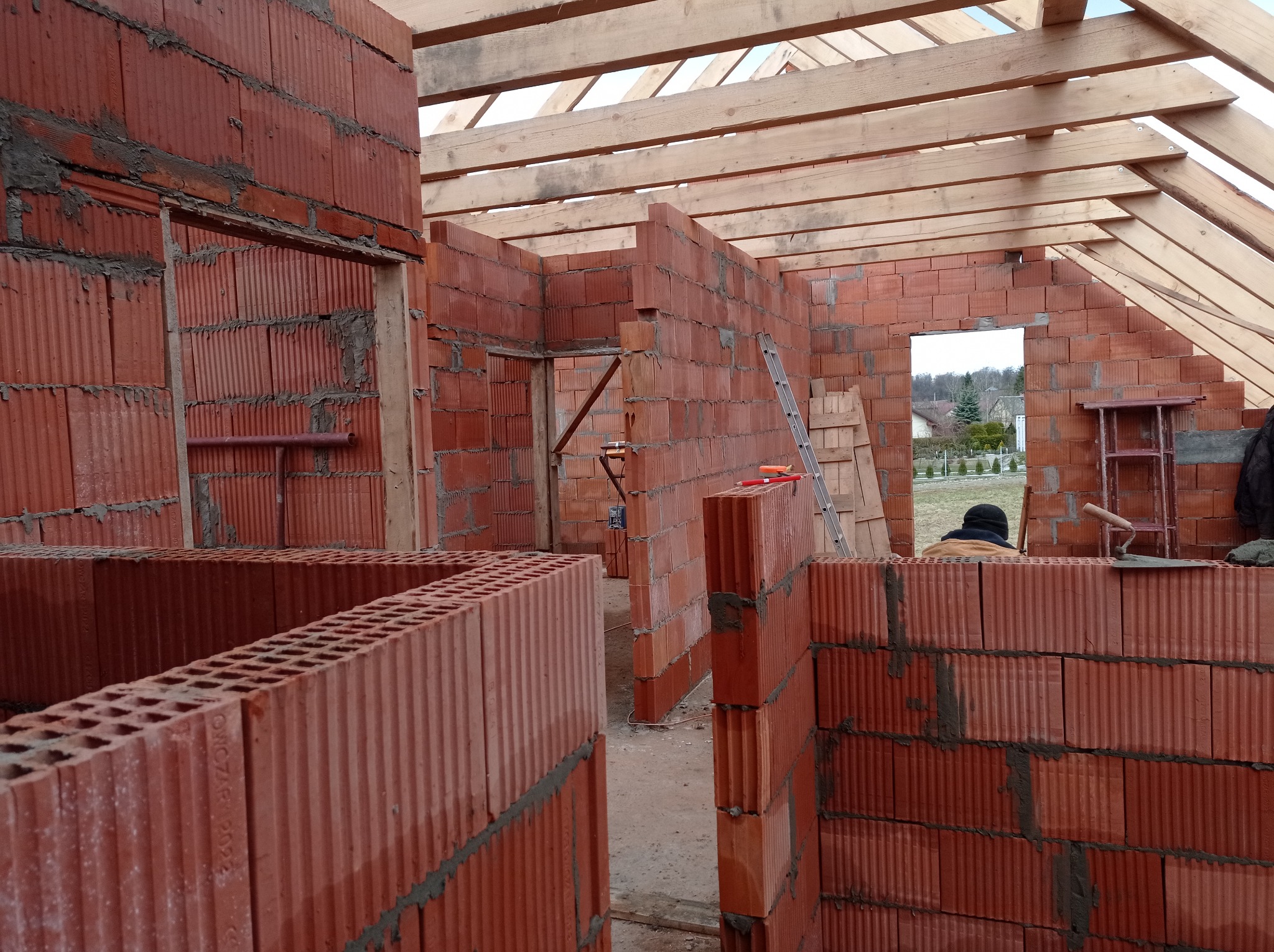 Wznoszenie ścian z czerwonej ceramiki poryzowanej na budowie domu, widoczne drewniane elementy konstrukcji dachu i drabina oparta o ścianę.