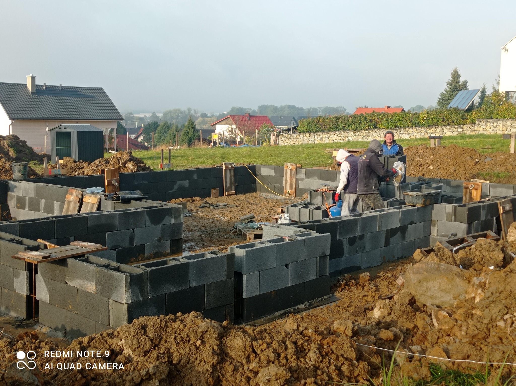 Budowa fundamentów domu z bloczków betonowych, widoczni trzej pracownicy, w tle zabudowa jednorodzinna i pochmurne niebo.