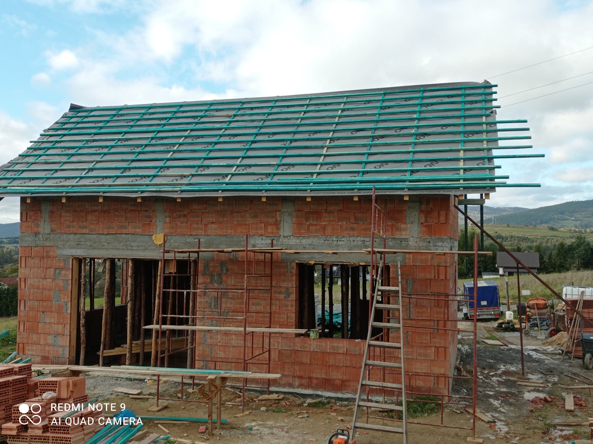 Surowy stan domu murowanego z czerwonej cegły, widoczne rusztowania, drabina oparta o ścianę, przygotowane materiały budowlane, w tle krajobraz z zabudowaniami.