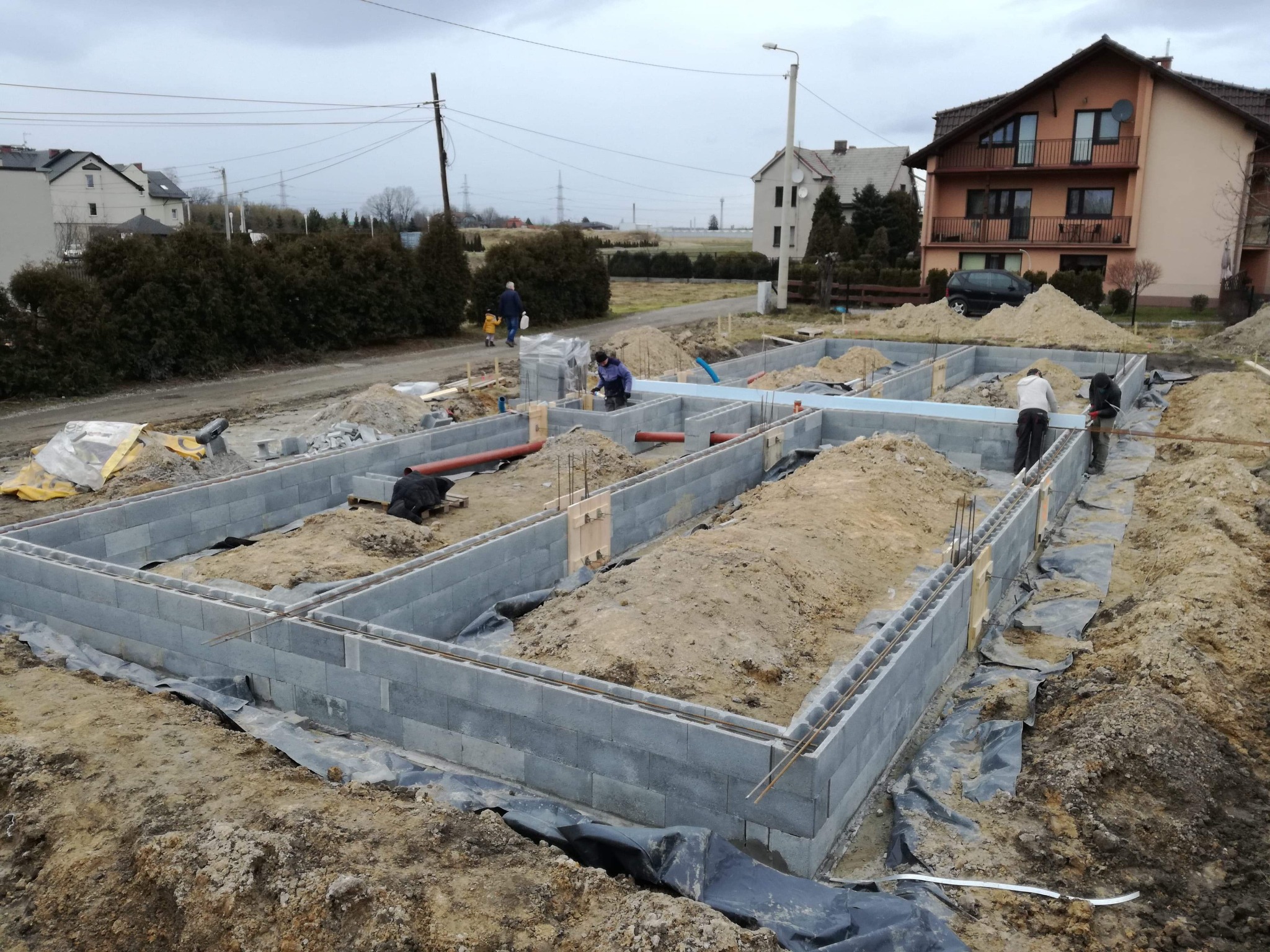 Budowa fundamentów domu z bloczków betonowych, widoczne zbrojenie, izolacja, rury kanalizacyjne oraz pracownicy na tle zabudowy jednorodzinnej w pochmurny dzień.