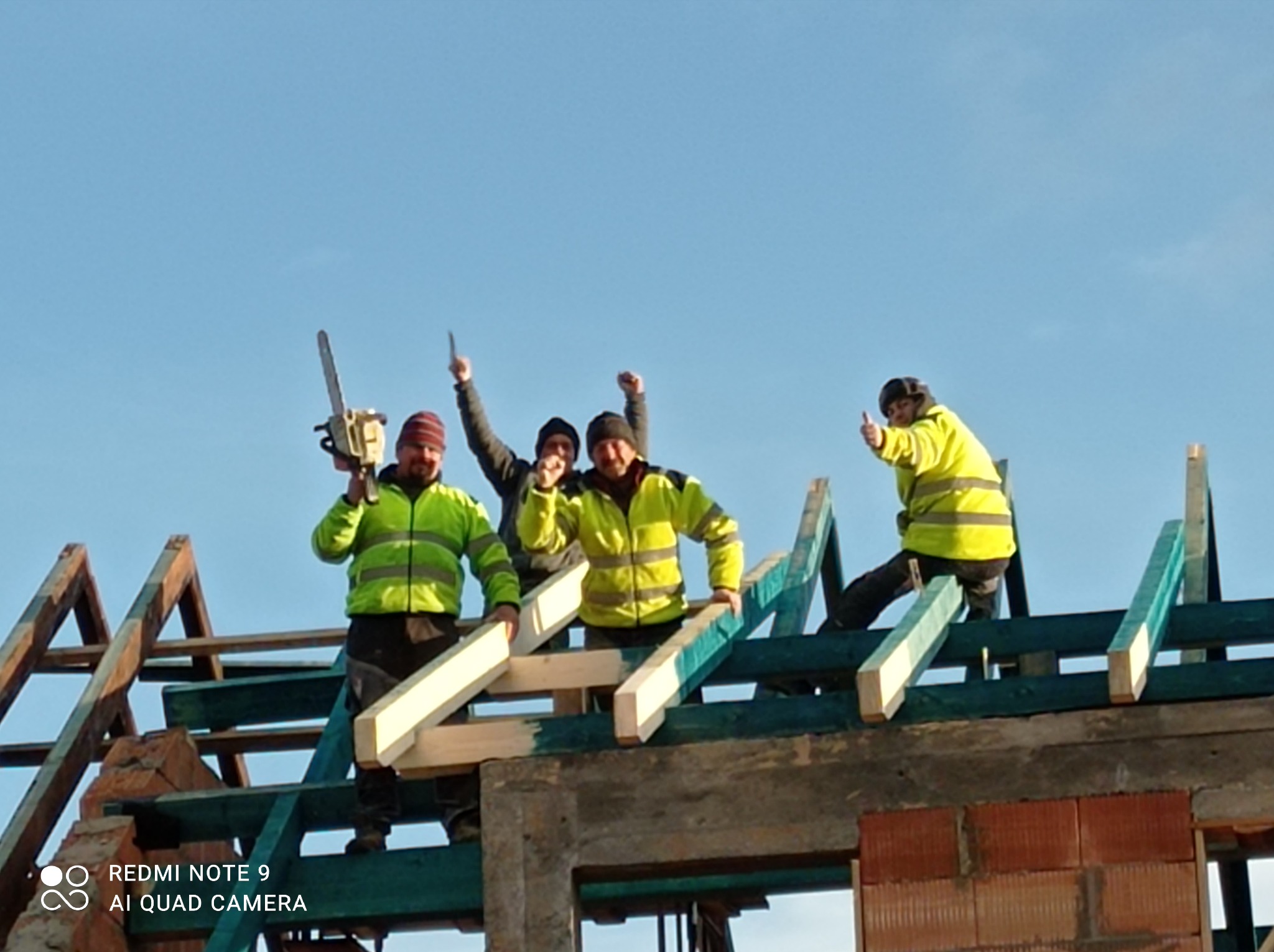 Grupa czterech roześmianych robotników w jaskrawych kurtkach na szczycie niedokończonej konstrukcji dachu z drewnianych belek, jeden z nich trzyma piłę łańcuchową, a drugi pokazuje kciuk w górę...