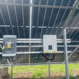 Panel fotowoltaiczny od spodu, widoczna skrzynka elektryczna z ostrzeżeniami i falownik na metalowej konstrukcji, trawa w tle.