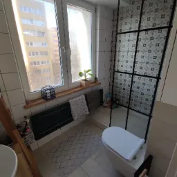 Nowoczesna łazienka z kabiną prysznicową typu walk-in z czarną ramą, białą toaletą i grzejnikiem drabinkowym pod oknem z widokiem na blok mieszkalny.