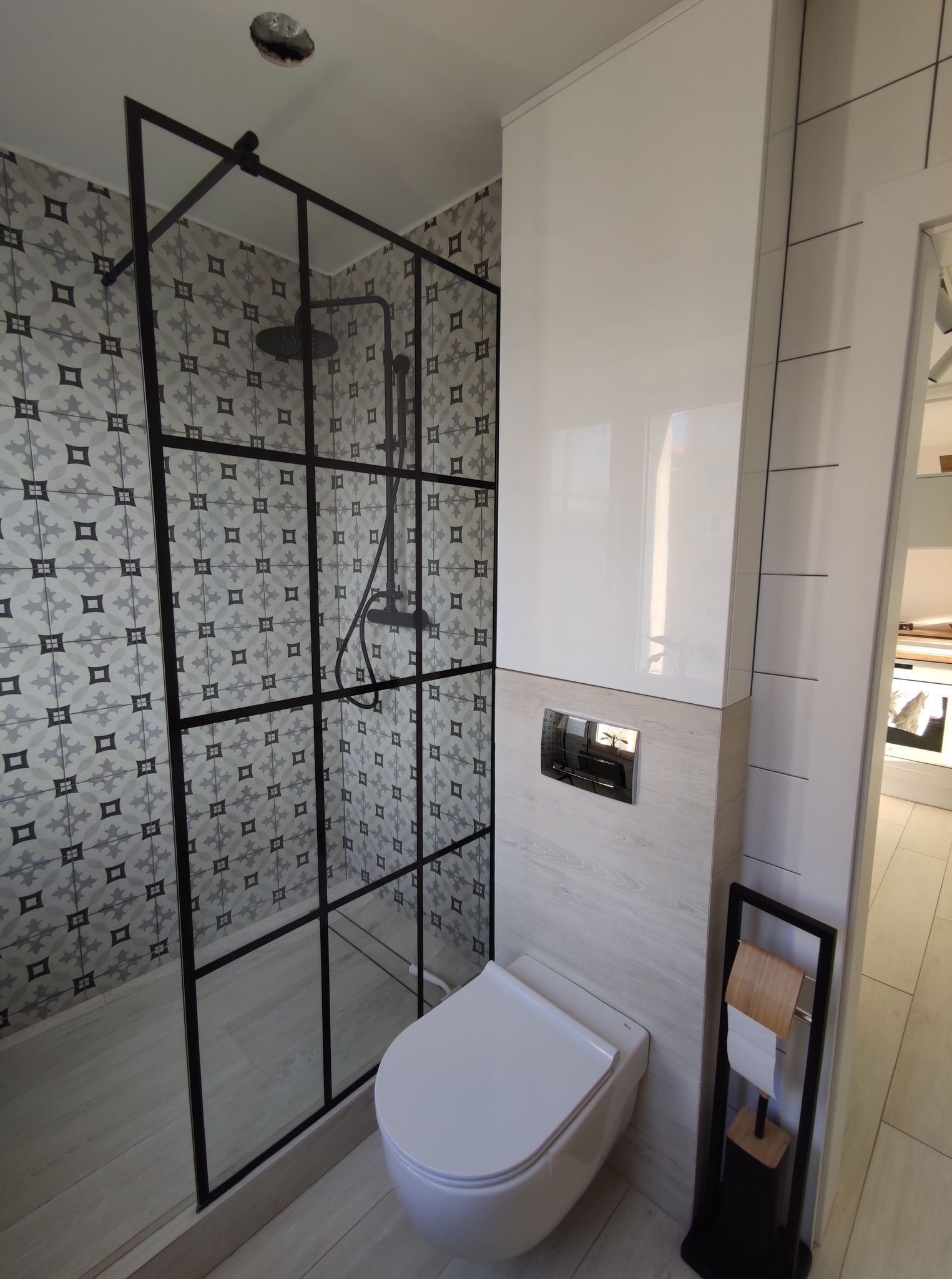 Nowoczesna łazienka z kabiną prysznicową typu walk-in z czarną ramą, wzorzystymi szarymi płytkami na ścianach w strefie prysznica, podwieszaną toaletą i minimalistycznym stojakiem na papier toaletowy
