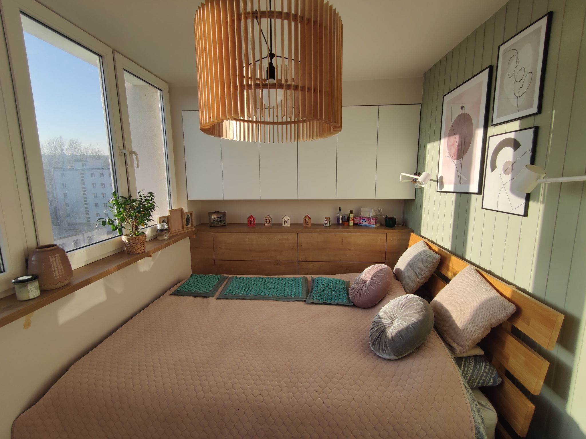 Jasna sypialnia z drewnianym zagłówkiem, pastelową pościelą i dekoracyjną lampą z drewnianych listewek. Na parapecie doniczka z zieloną rośliną i ozdobne przedmioty. Ściany w odcieniach bieli...