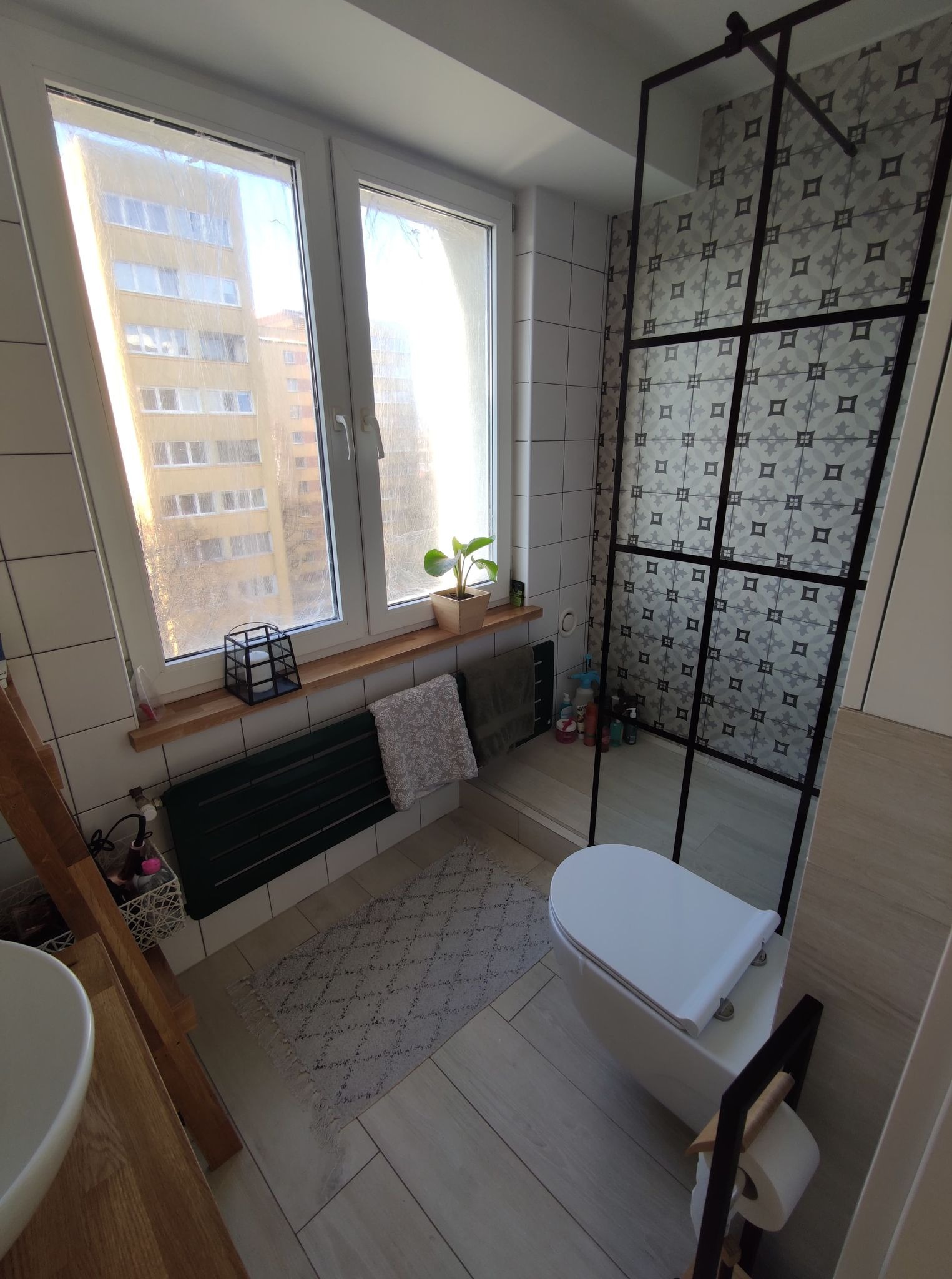 Nowoczesna łazienka z kabiną prysznicową typu walk-in z czarną ramą, białą toaletą i grzejnikiem drabinkowym pod oknem z widokiem na blok mieszkalny.