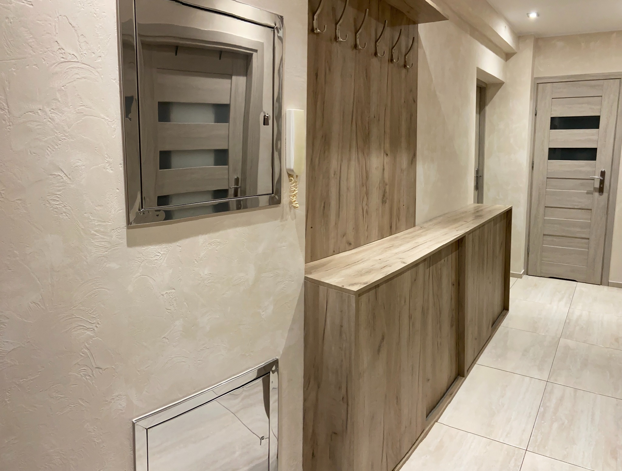 Wykończony przedpokój z beżowymi ścianami o fakturze baranka, zabudową z drewnopodobnej płyty z wieszakami i szafkami, lustrem w metalowej ramie oraz drzwiami z poziomymi przeszkleniami.