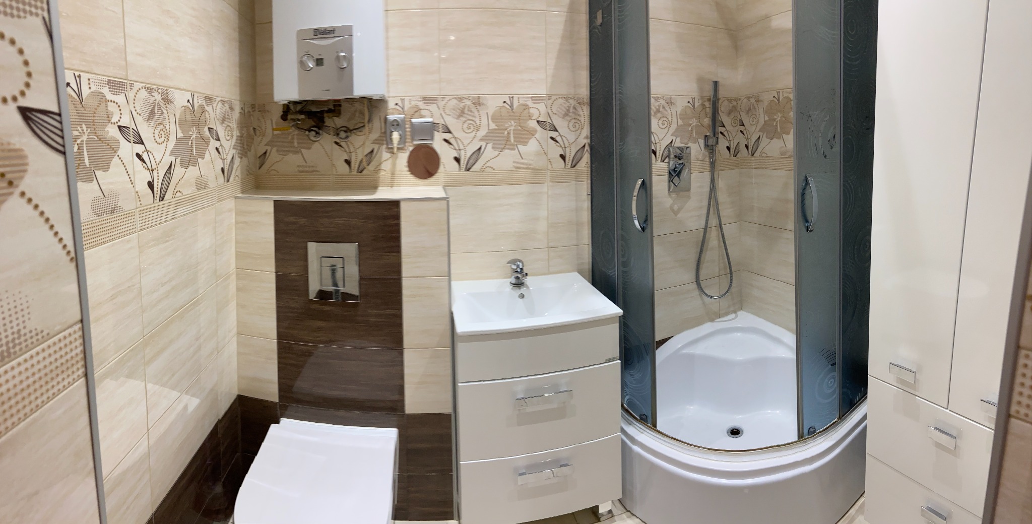 Wykończona łazienka z beżowymi płytkami z motywem kwiatowym, białą szafką z umywalką, kabiną prysznicową z grafitowymi drzwiami i białym brodzikiem oraz podgrzewaczem wody na ścianie.